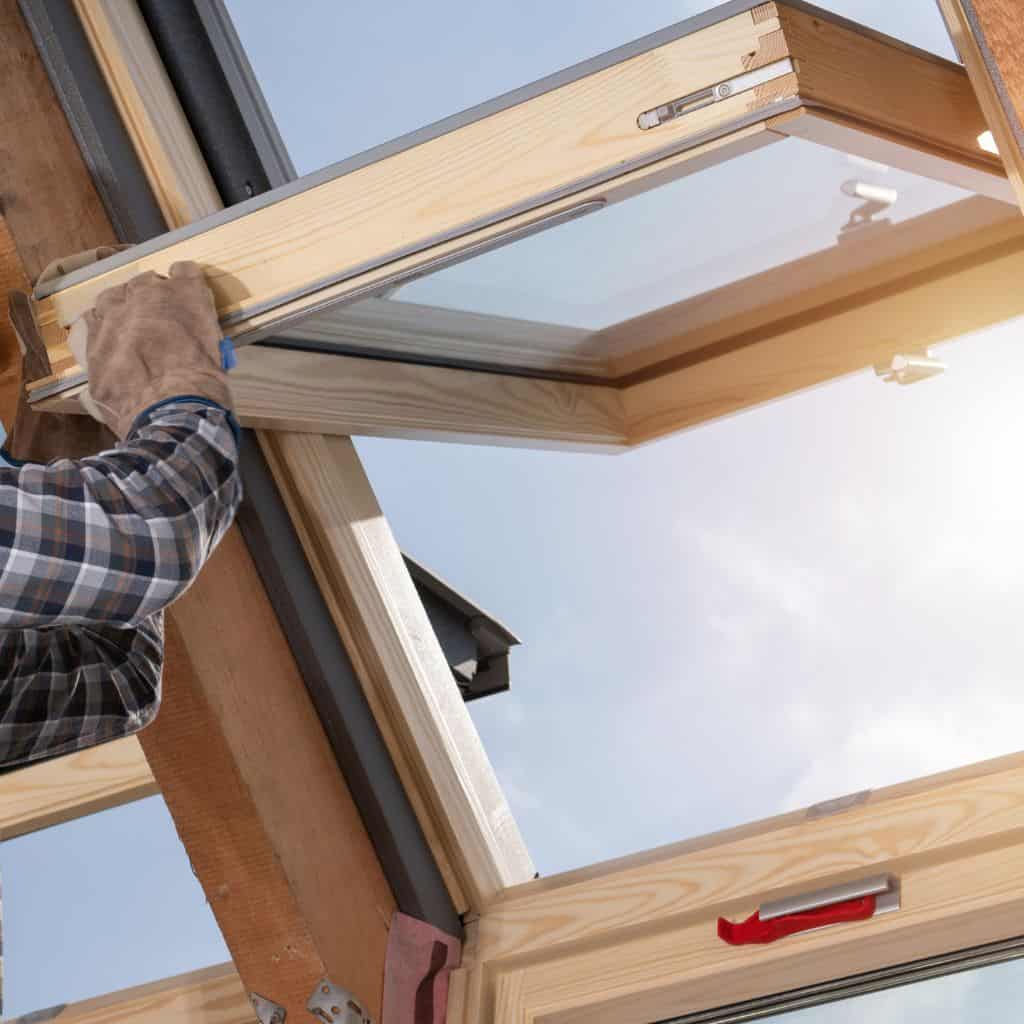 Worker fitting a timber-framed roof window into a pitched roof opening against a bright sky.