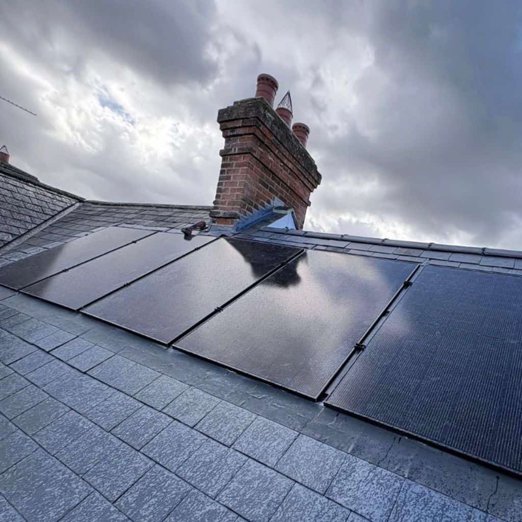 Black solar panels fitted across a grey slate roof beside a tall red brick chimney under a cloudy sky.