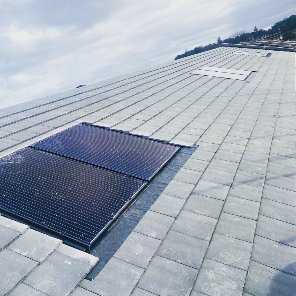 Black solar PV panels being installed on a grey tiled pitched roof under a cloudy sky.