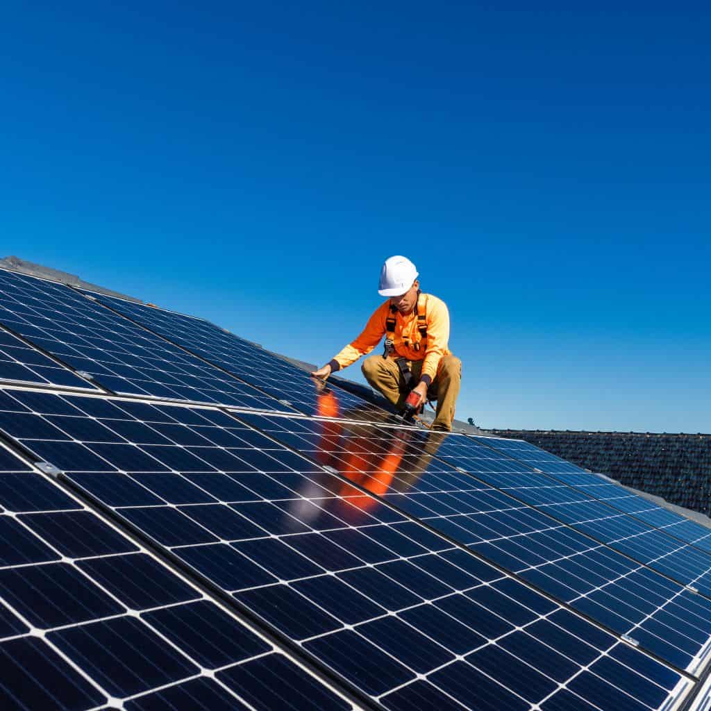 Worker in orange high-visibility clothing and white hard hat fitting solar panels on a sunlit roof under blue sky.
