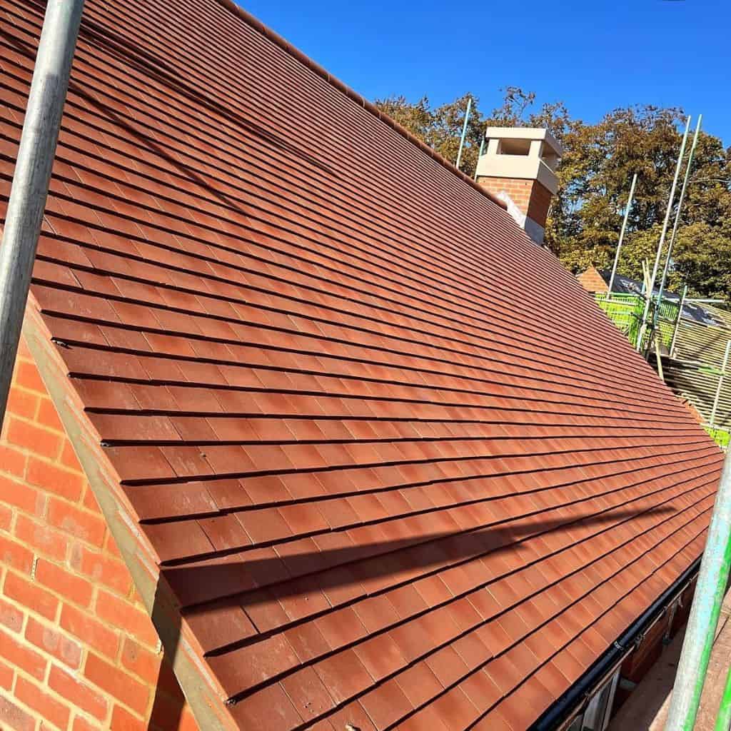Newly laid red clay tile pitched roof on a house, with scaffolding poles and a brick chimney against a blue sky.