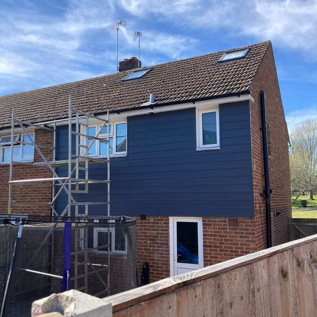 House gable clad in dark grey horizontal wall cladding above brickwork, with scaffolding and white windows.