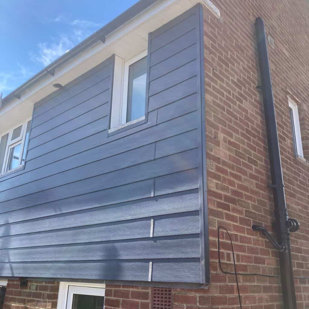 Roofer in black fitting grey weatherboard cladding to a dormer with a dark window, working from scaffolding on a roof.