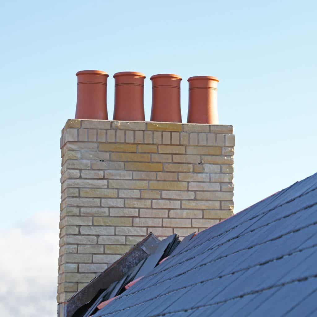 Cream brick chimney stack with four red terracotta pots above a dark slate roof against a blue sky.