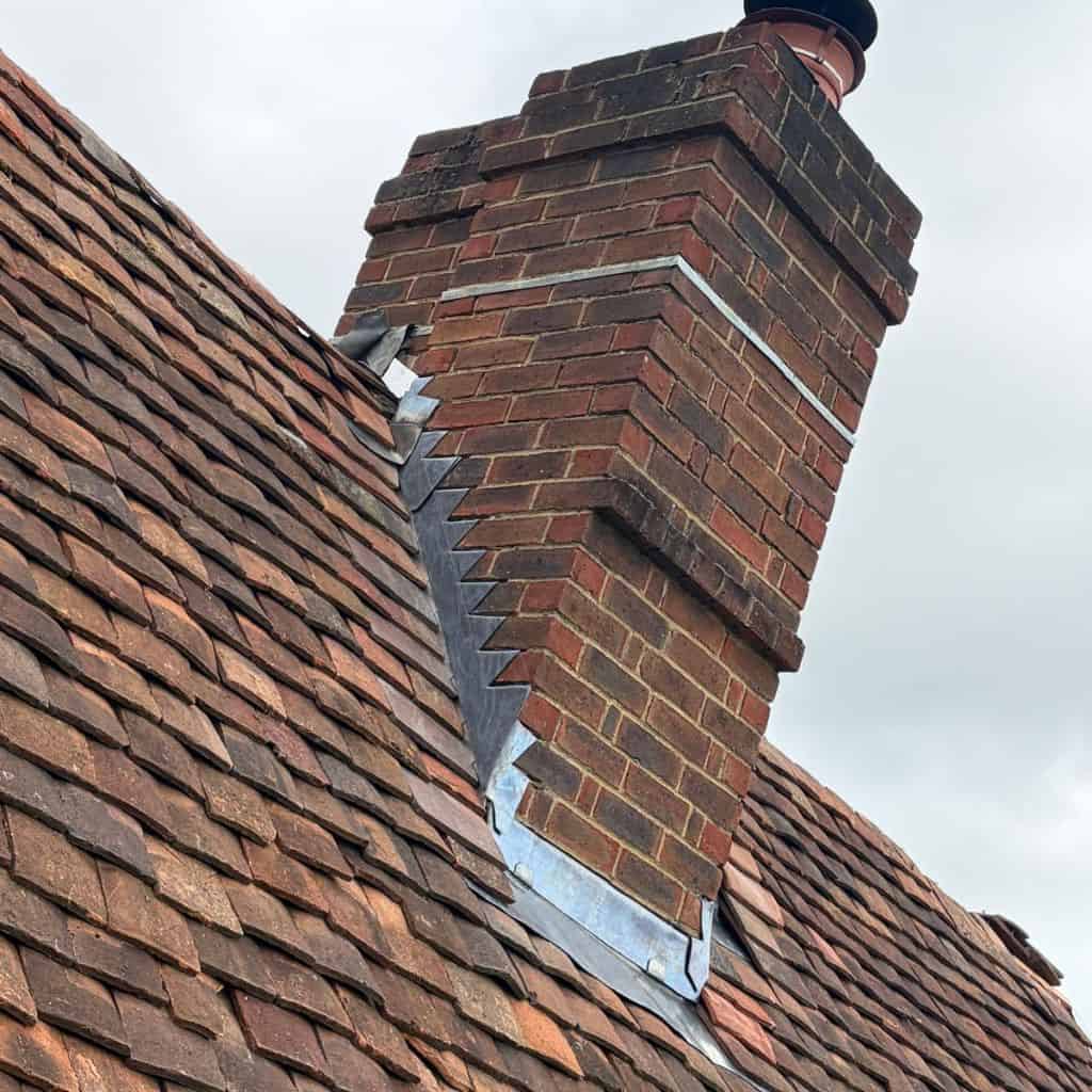 Brick chimney stack with new stepped lead flashing sealing the junction with an old red clay tiled roof under a grey sky.