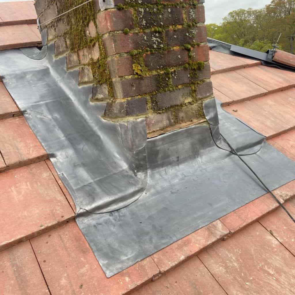 Fresh grey lead flashing dressed around a moss-covered brick chimney where it meets a red clay tile roof.