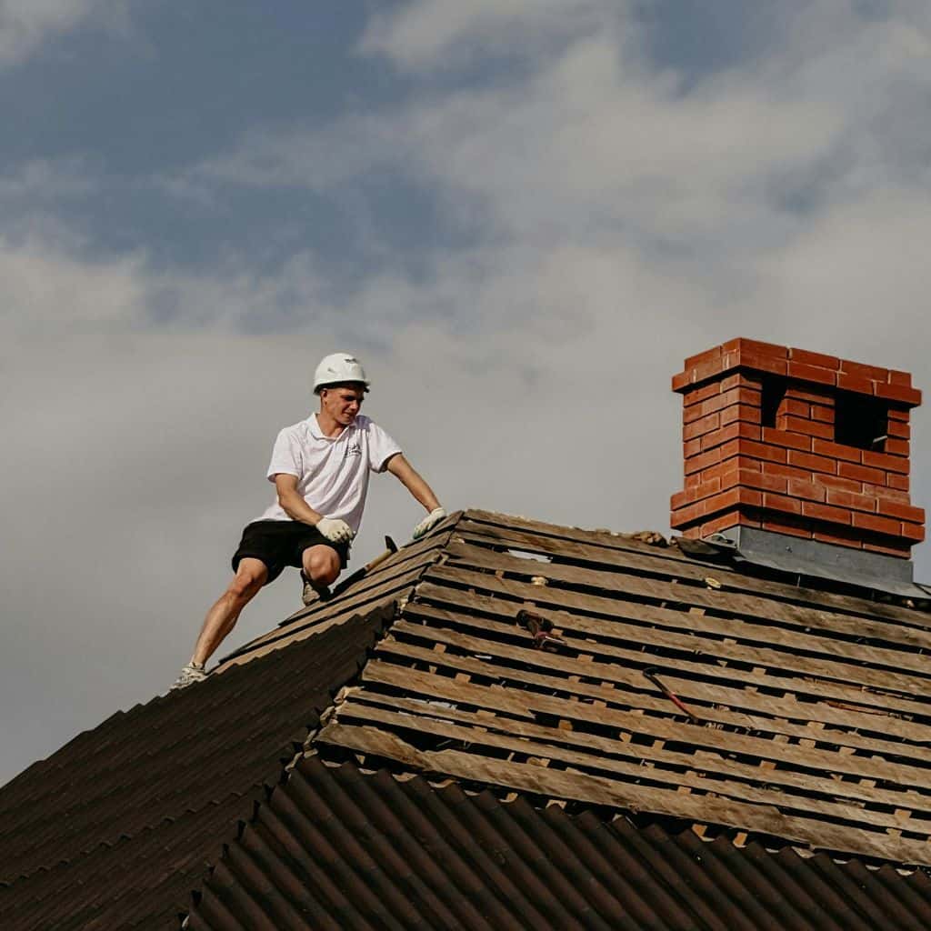 Roofer in a white hard hat on a stripped roof beside a tall red brick chimney stack against a cloudy sky.