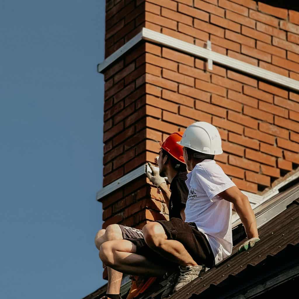 Two workers in hard hats sitting on a roof inspecting the brickwork of a tall red brick chimney stack.