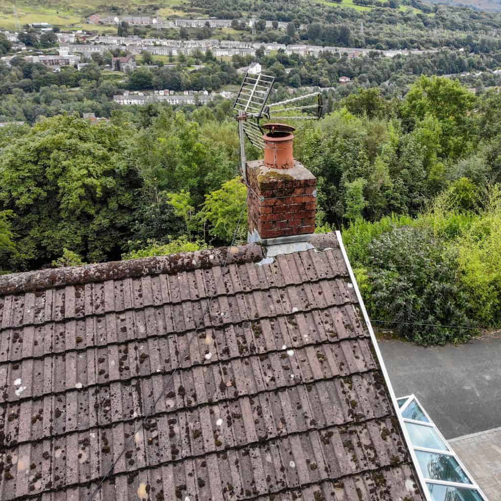 Aerial view of a moss-stained brick chimney with terracotta pot and aerial on a weathered tile roof above a valley town.