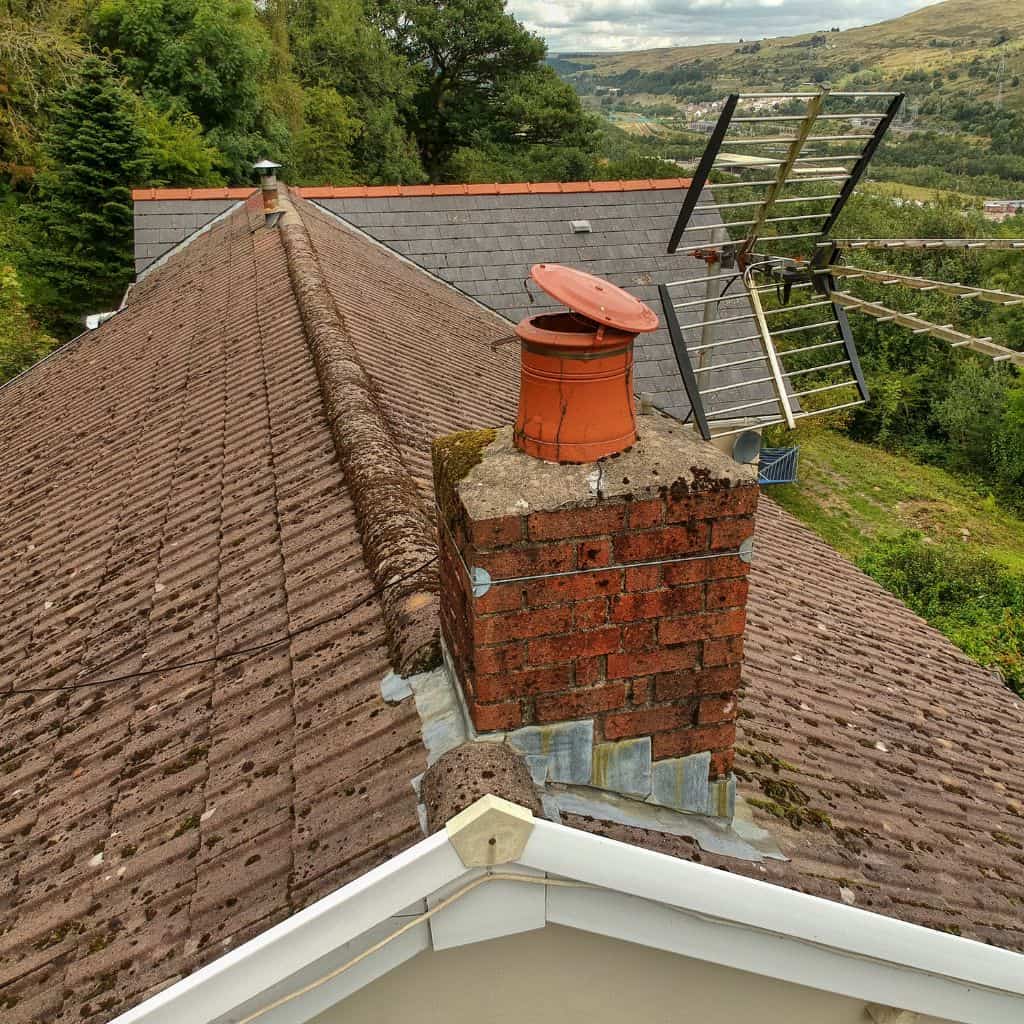 Aerial view of a brick chimney with a terracotta pot and hinged cowl on an aged slate roof above a hillside.