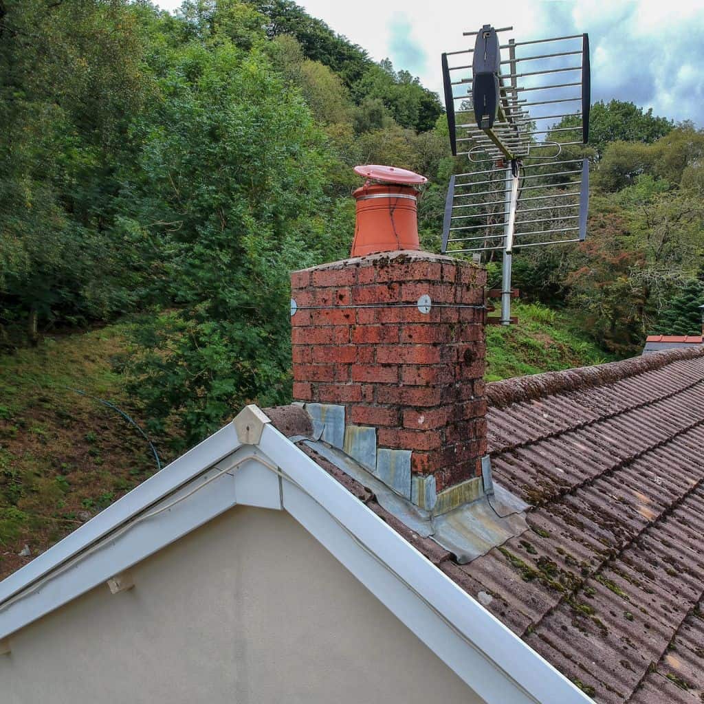 Red brick chimney with a terracotta pot and aged lead flashing at its base, beside a mossy concrete tiled roof.