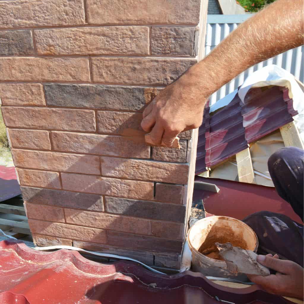 Roofer's hand pressing fresh mortar into the brickwork joints of a chimney stack above a red metal tile roof.