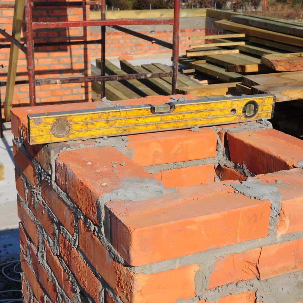 Red brick chimney being rebuilt with fresh mortar, a yellow spirit level resting on top, scaffolding and timber behind.