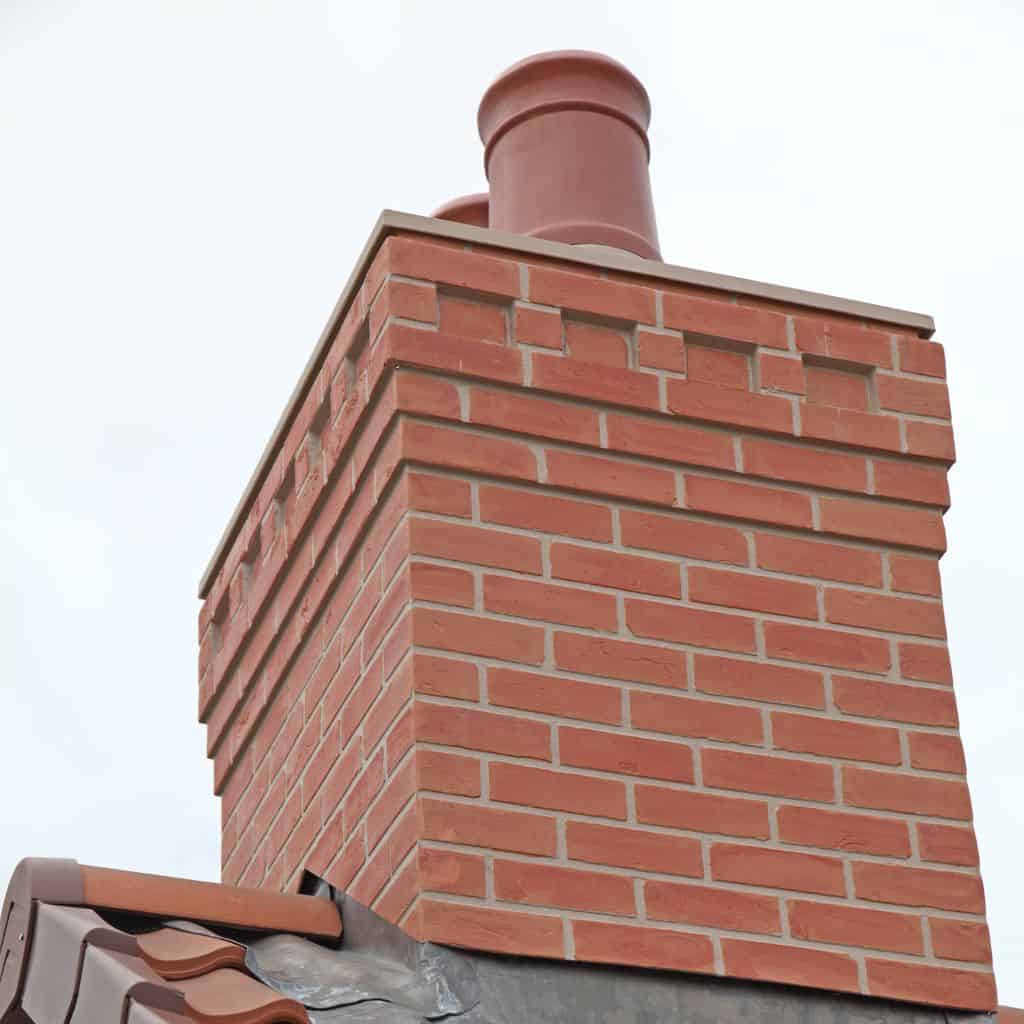Newly rebuilt red brick chimney stack with corbelled brick courses and a terracotta pot against a pale sky.