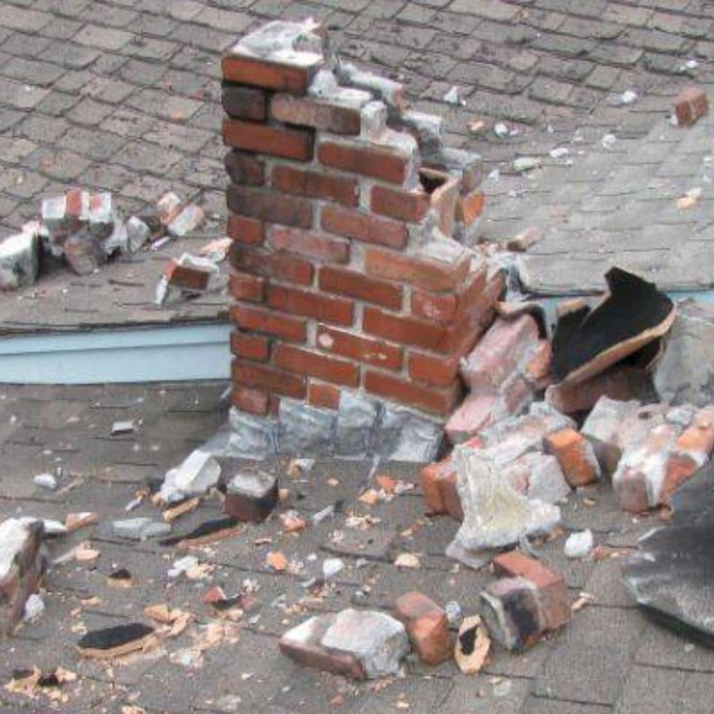 Partly demolished red brick chimney stack on a shingle roof with loose bricks and rubble scattered around it.