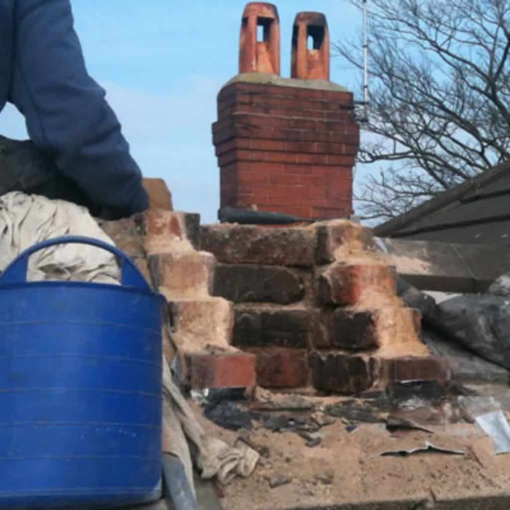 Roofer kneeling beside a partly demolished brick chimney with a blue rubble bucket, twin pots on a stack behind.