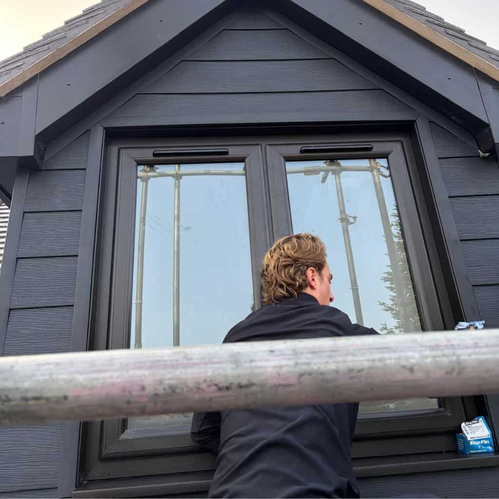 Worker in black seen from behind on scaffolding cleaning a uPVC window set into dark grey weatherboard cladding on a gable.