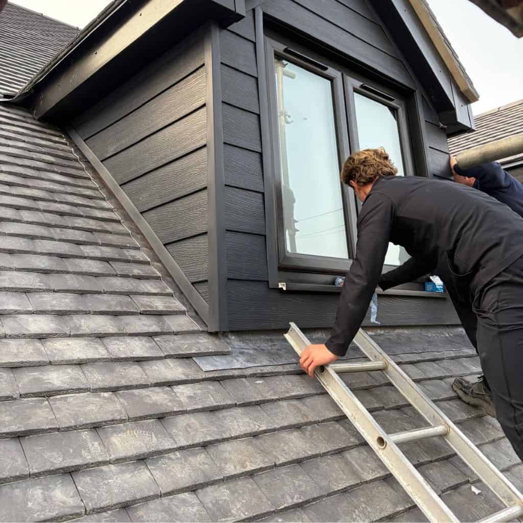 Two roofers handling a ladder beside a dormer clad in dark cladding above a grey slate roof.