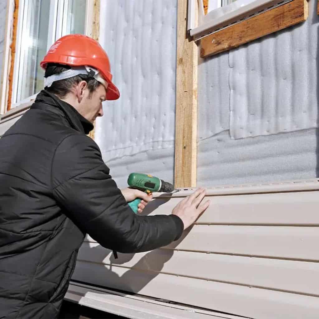 Worker in a red hard hat using a drill to fix beige horizontal wall cladding to a timber-framed wall.