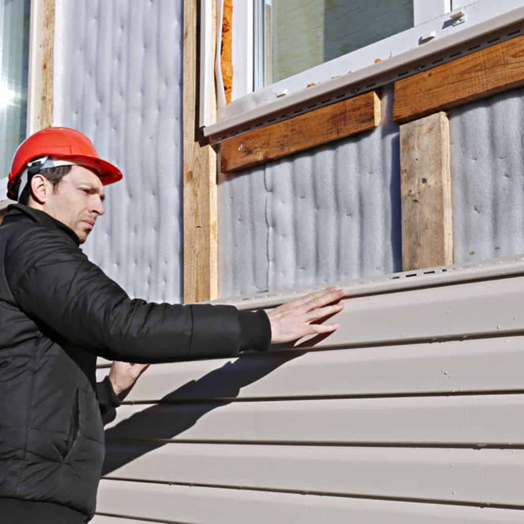 Man in a black jacket and red hard hat fitting beige horizontal cladding boards to a house wall in the sun.