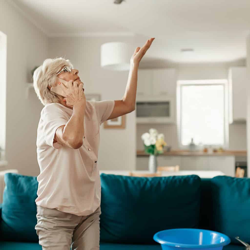 Older woman on the phone reaching up towards a ceiling with a blue bucket on the sofa to catch a leak.