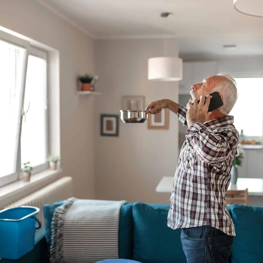 Older man on the phone in a living room, holding a pan to catch a ceiling leak, with a bucket by the sofa.