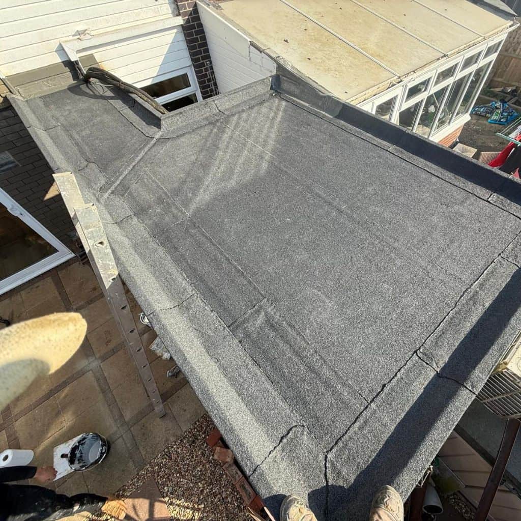 Completed grey felt flat roof on a rear extension beside a conservatory, with a roofer's feet visible at the edge.