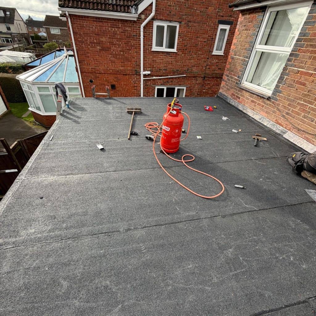 Worker in a red cap laying black felt over a flat roof, with rolls and primer beside fresh timber decking.