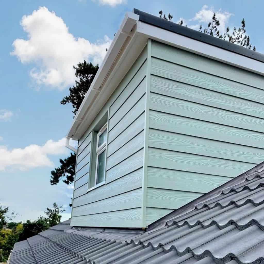 Pale green fibre cement weatherboard cladding on an extension above a grey tile roof, against a blue sky.