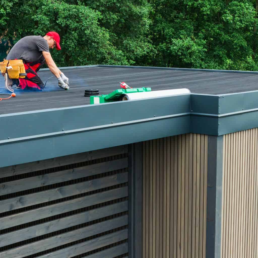 Roofer in a red cap kneeling on a dark EPDM flat roof using a torch tool, beside timber-clad walls and trees.