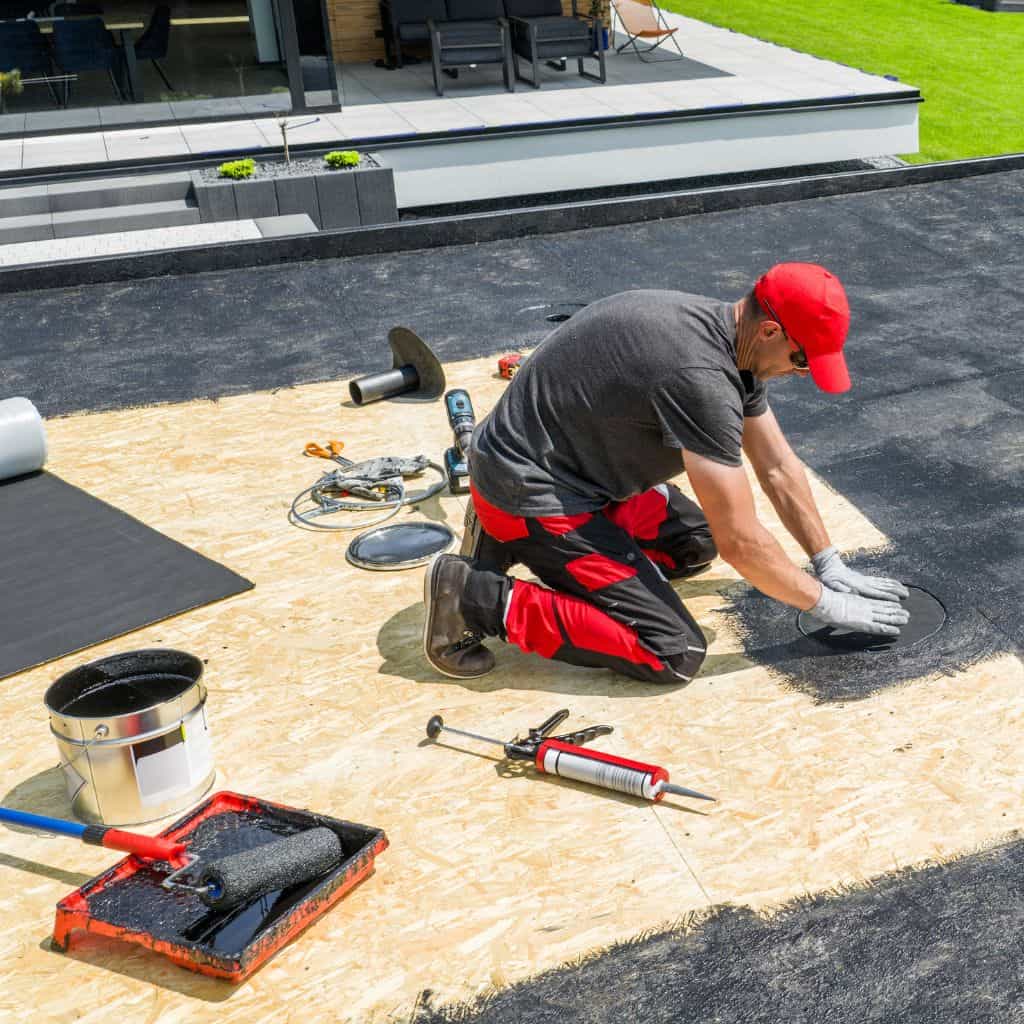 Roofer in a red cap kneeling on a flat roof bonding black membrane over timber boards, with tools and a paint roller nearby.