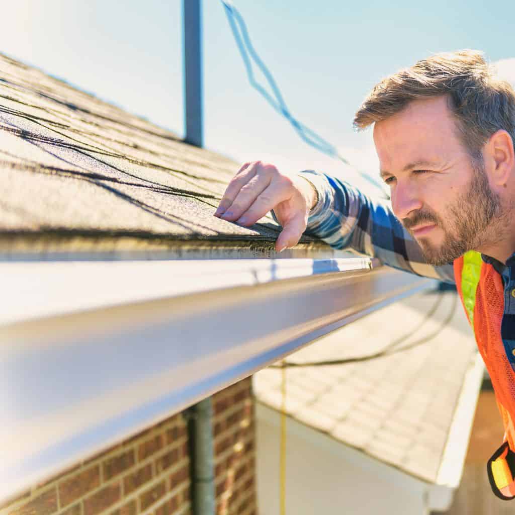 Man in a hi-vis vest leaning over a roof edge to inspect the guttering and shingle roof in bright sunshine.