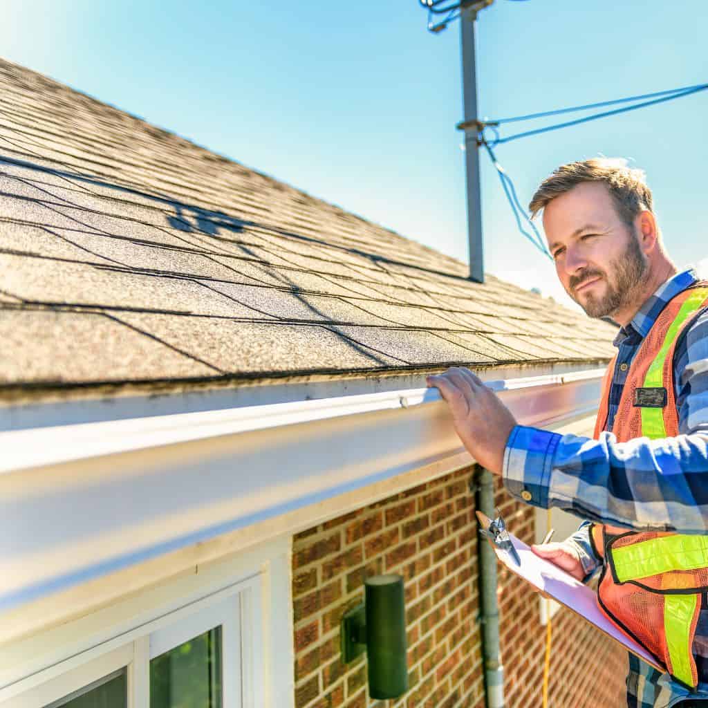 Roofer in a hi-vis vest inspecting a white gutter at the eaves of an asphalt shingle roof, holding a clipboard.