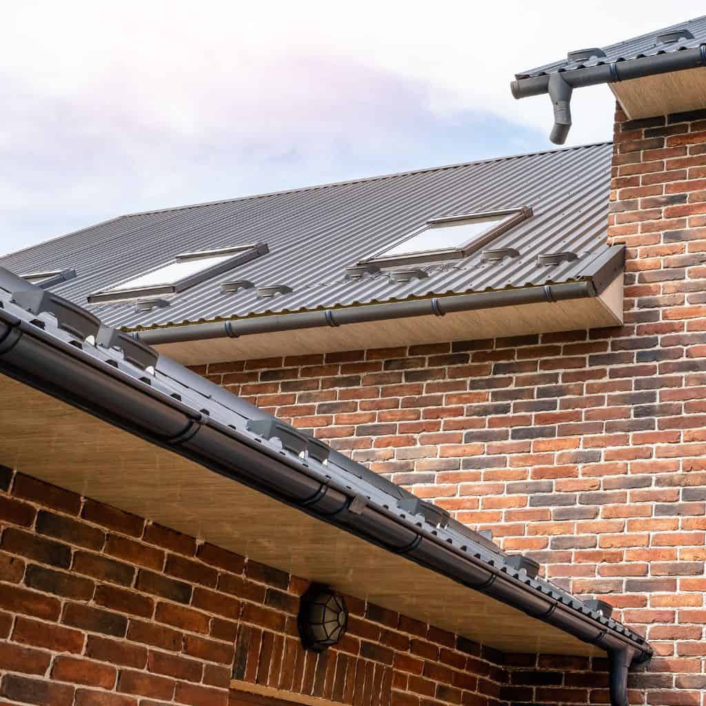 Black guttering and downpipes fitted along the eaves of a brick house with a corrugated roof and rooflights.