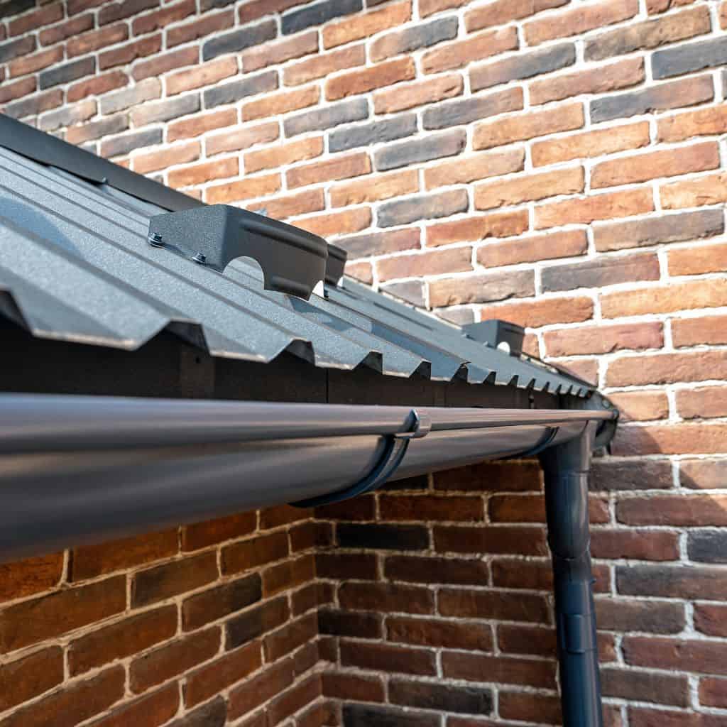 Close-up of a dark metal roof edge with eaves clips draining into a grey gutter against a brick wall.