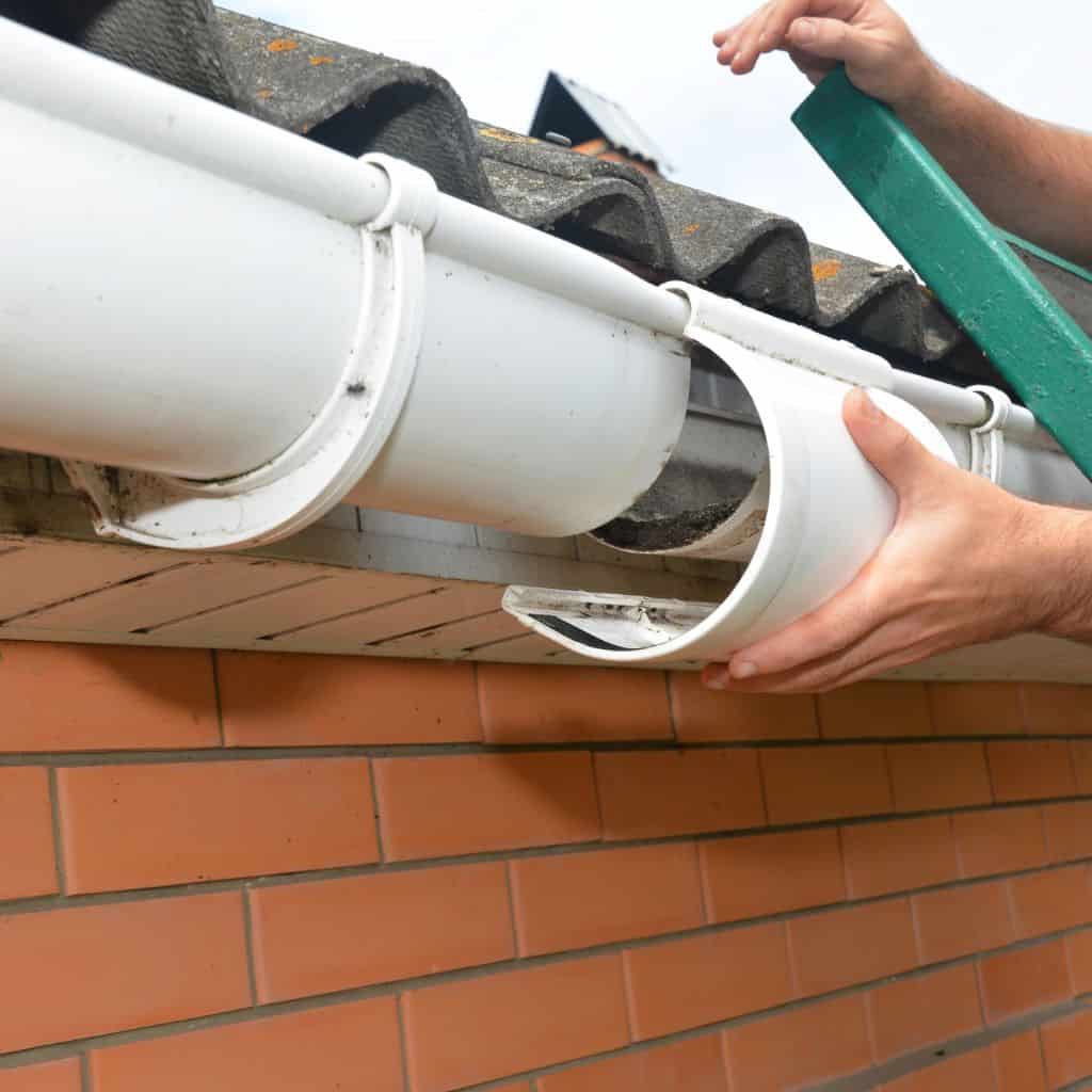 Hands removing a section of white uPVC half-round guttering from a roof edge above red brickwork.