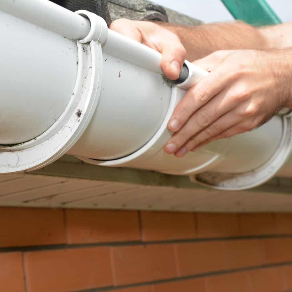 Hands fitting a white PVC gutter onto fascia above an orange brick wall, joining two gutter sections.