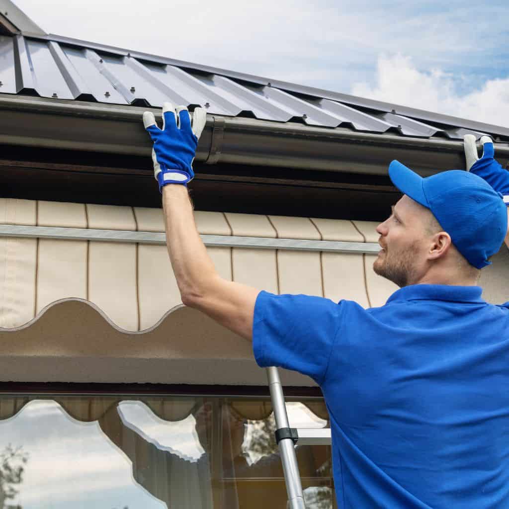 Roofer in a blue shirt and gloves fitting guttering beneath a dark metal roof edge from a ladder.