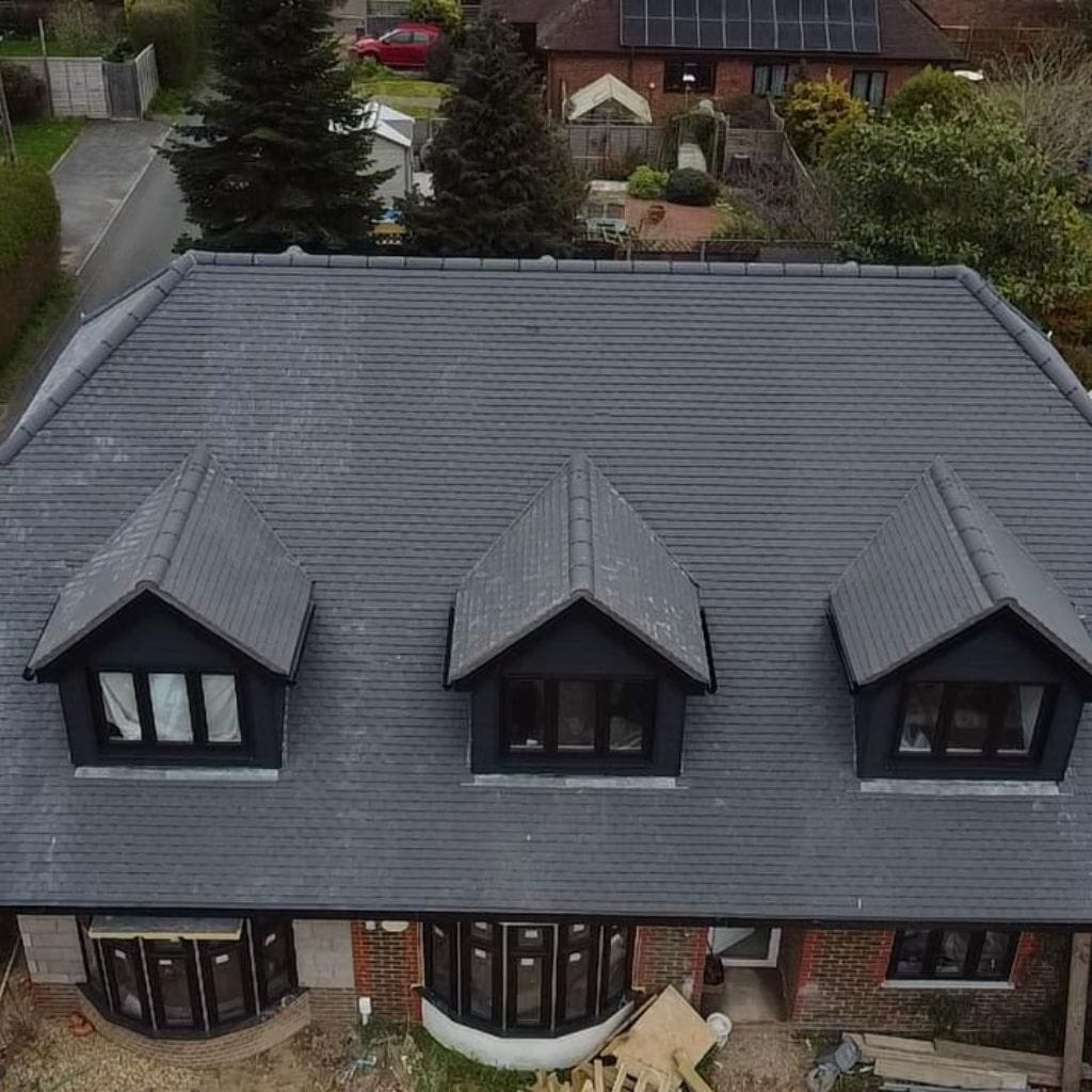 Aerial view of a house with a new grey tiled roof and three matching dark dormer windows, surrounded by gardens.