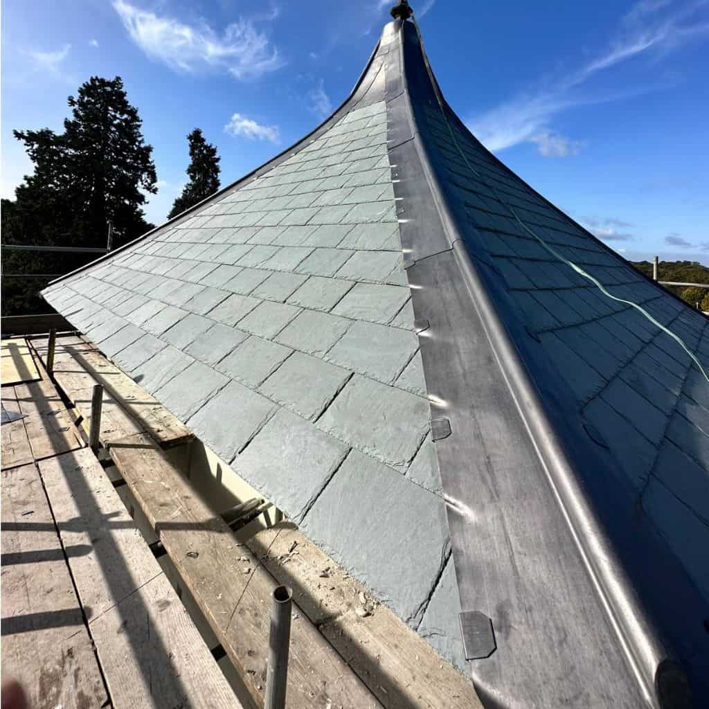 Curved conical roof being clad in grey slates with bright lead detailing, viewed from scaffold boards against a blue sky.