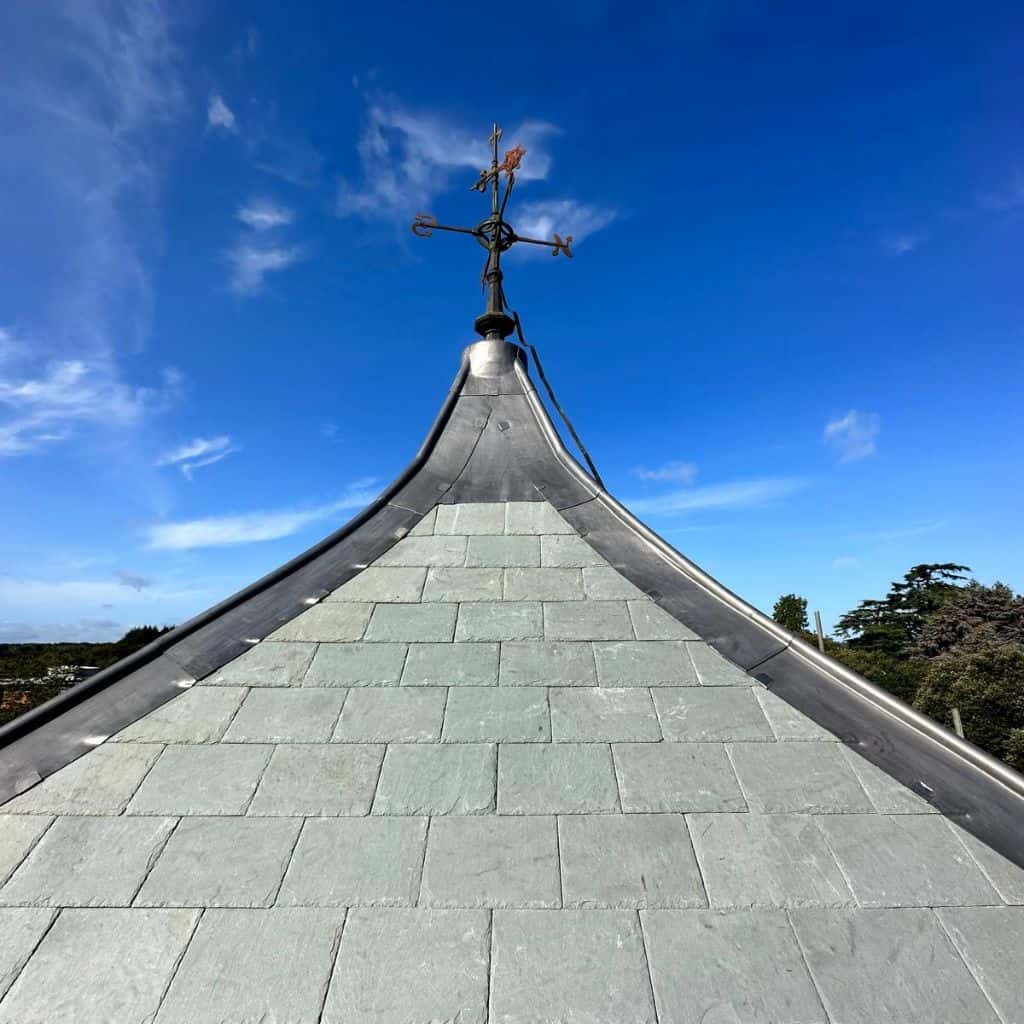 Conical grey slate spire topped with an ornate metal cross and weathervane against a deep blue sky.