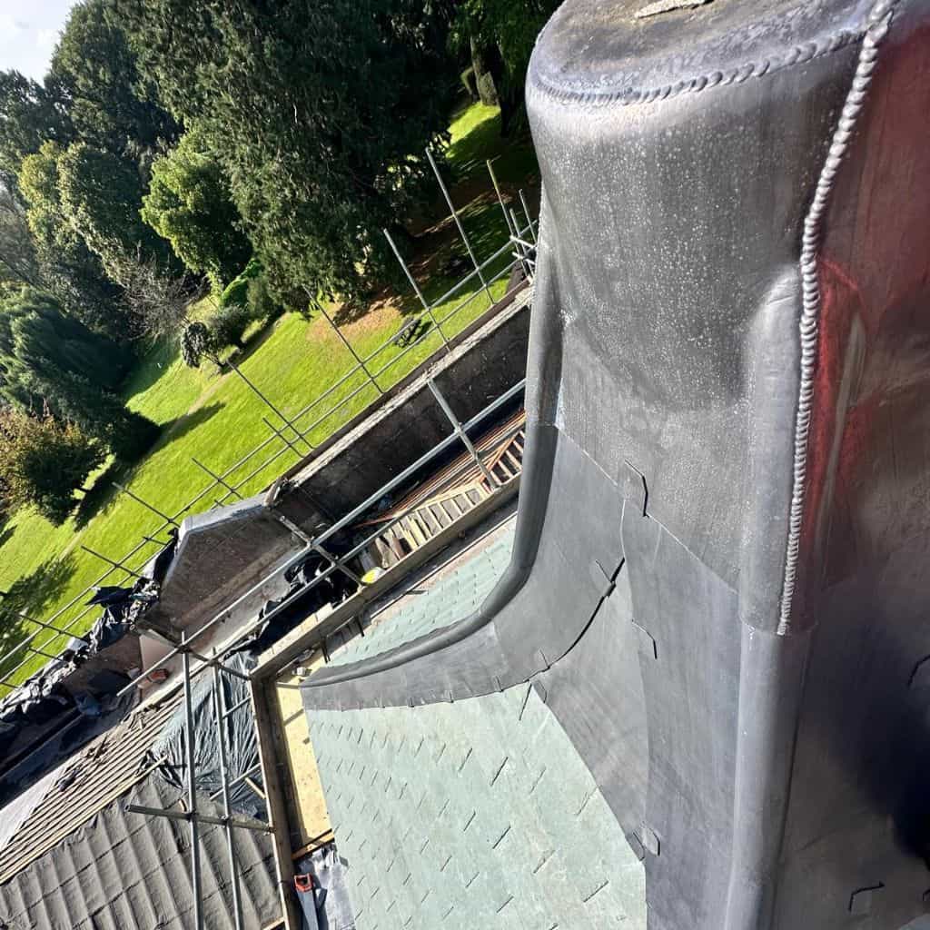 Newly dressed grey lead flashing wrapping around a roof upstand on a slate roof, with scaffolding and a green lawn beyond.