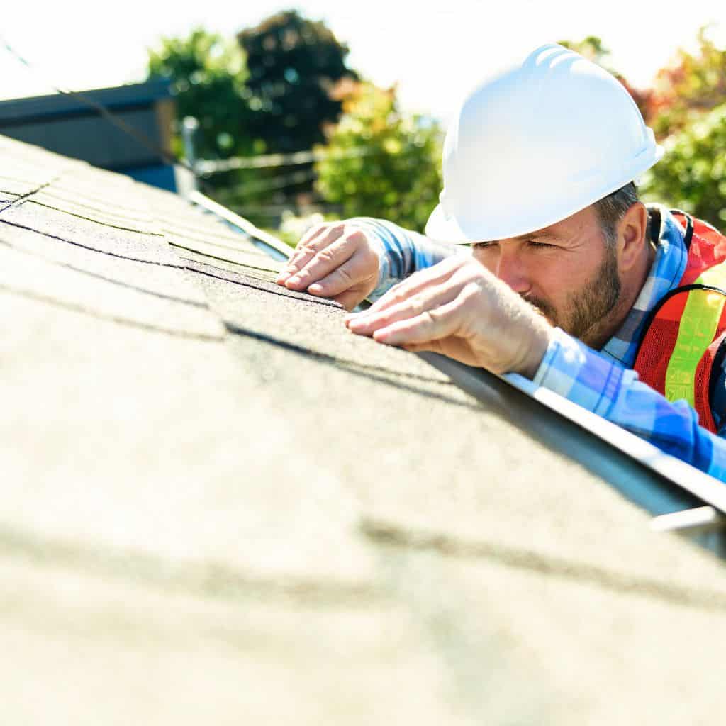 Inspector in a hard hat and hi-vis vest leaning over a roof ridge to examine shingles up close.