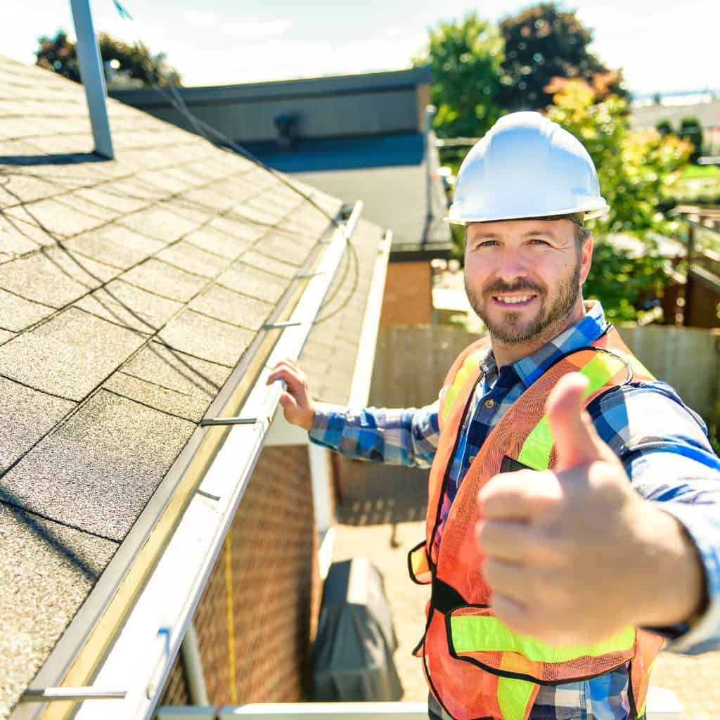 Smiling worker in a hard hat and hi-vis vest giving a thumbs-up beside a shingle roof and gutter during an inspection.