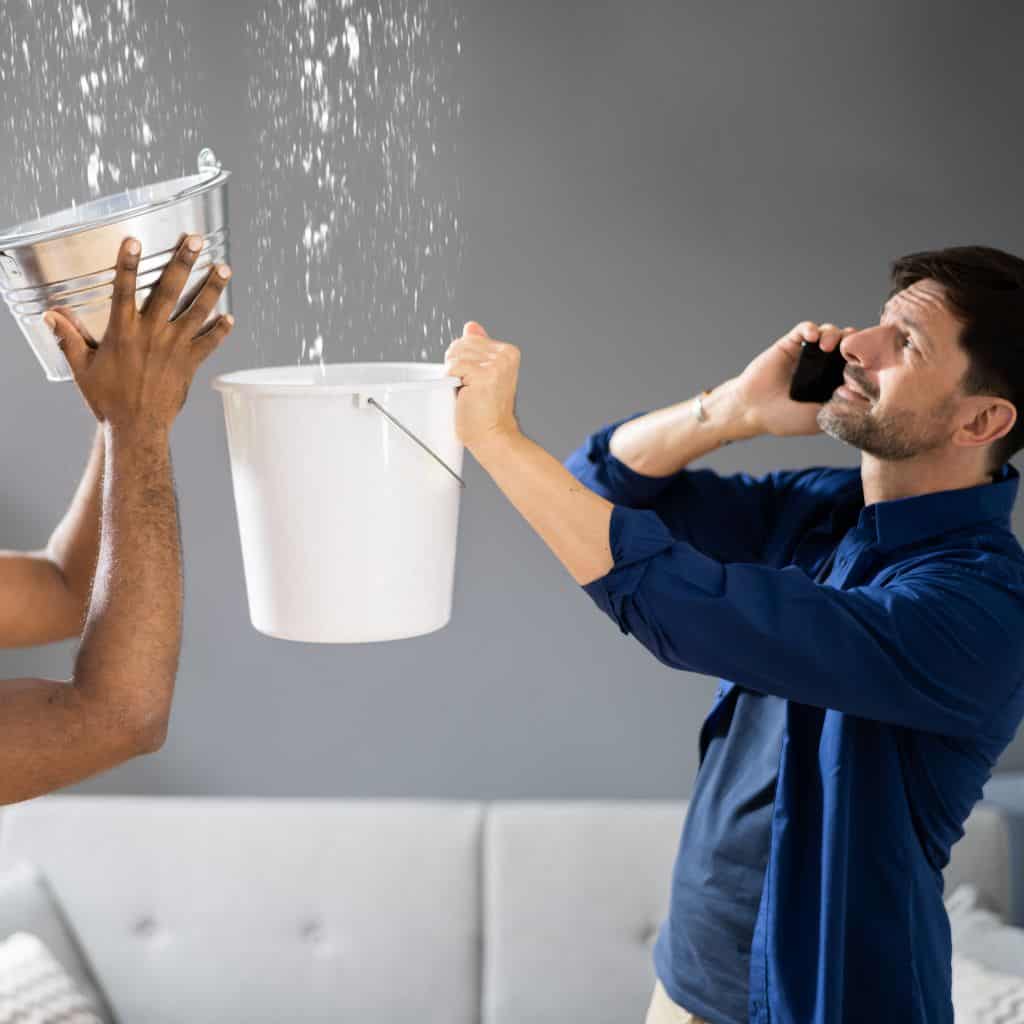 Two people catching dripping water in buckets indoors while one makes a phone call beneath a leaking ceiling.