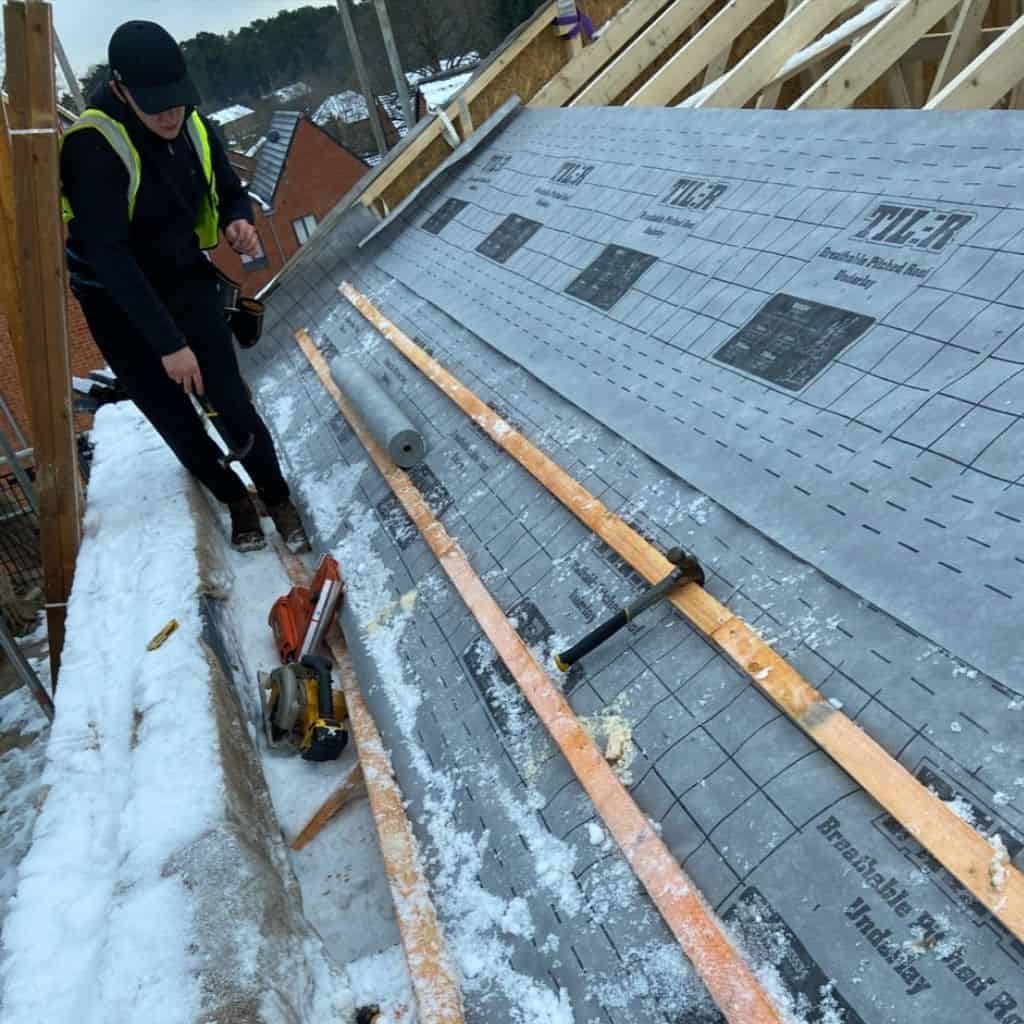 Roofer in hi-vis cutting timber battens on a snow-dusted roof covered with grey breathable underlay and tools.