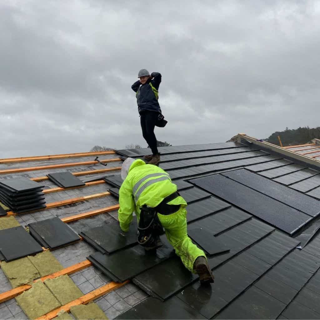 Two roofers on a pitched roof laying dark grey slates over battens and membrane on an overcast, windy day.