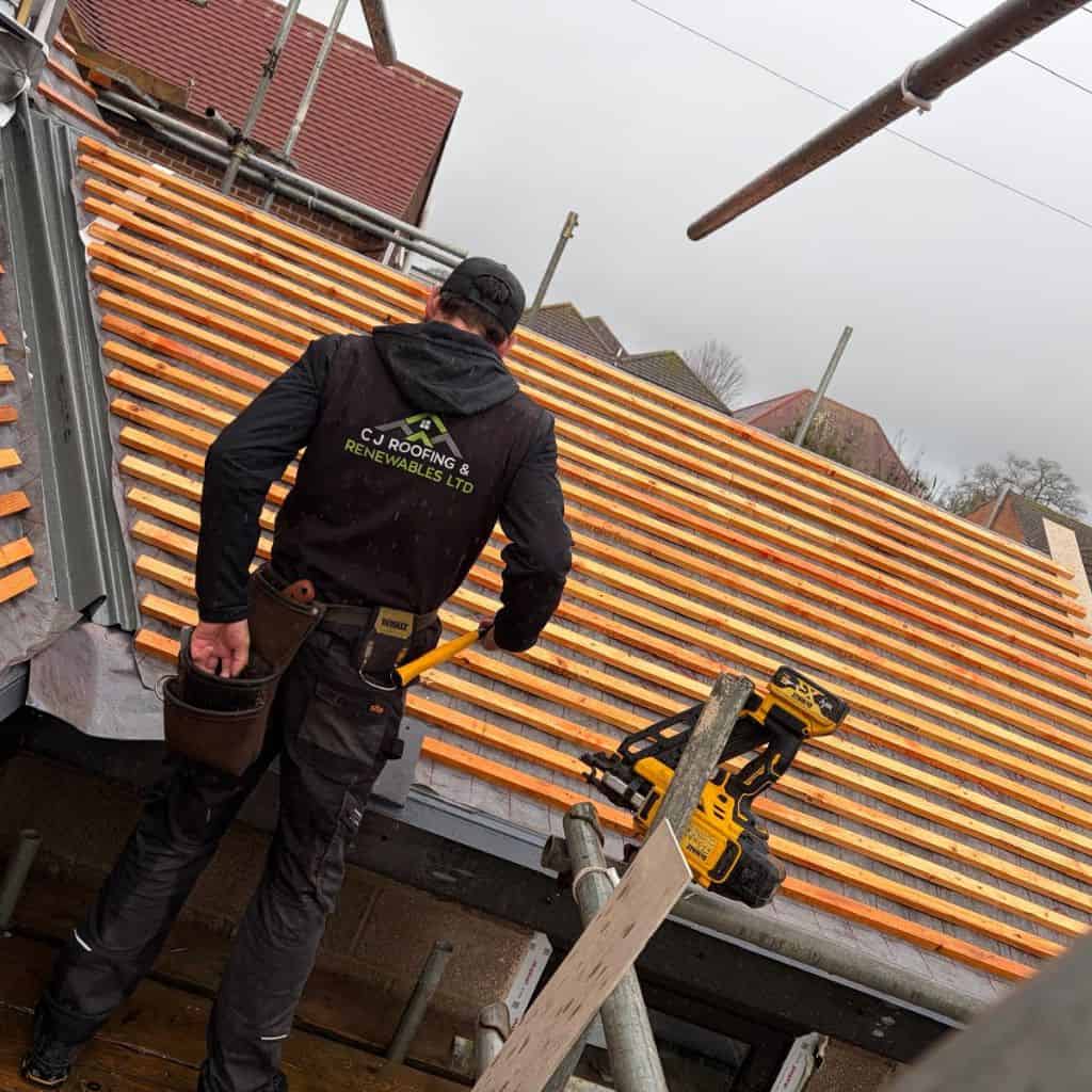 C J Roofing & Renewables worker seen from behind fitting timber battens across a stripped roof, with a nail gun nearby.