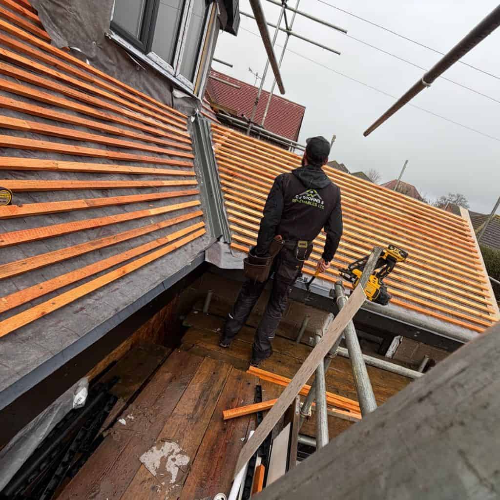 Roofer in branded workwear on scaffolding battening a pitched roof, with tools and a nail gun beside him.