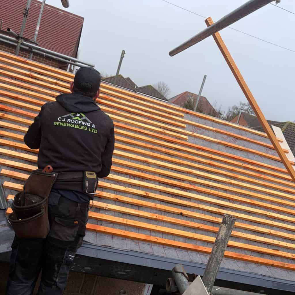 Roofer in a C J Roofing & Renewables hoodie fixing timber battens to a stripped roof, with a tool belt and scaffolding.